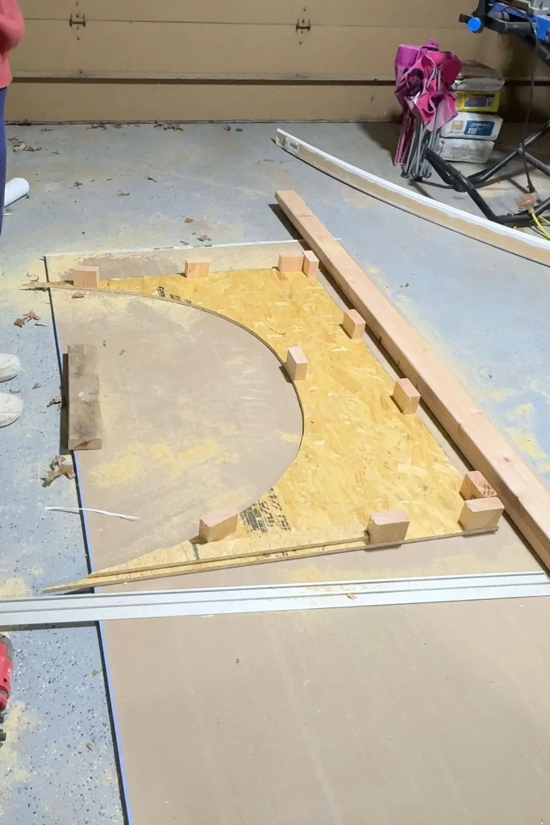 A partially completed wooden arch made from OSB sheeting rests on the garage floor. The arch is supported by small wood blocks, surrounded by sawdust and tools.
